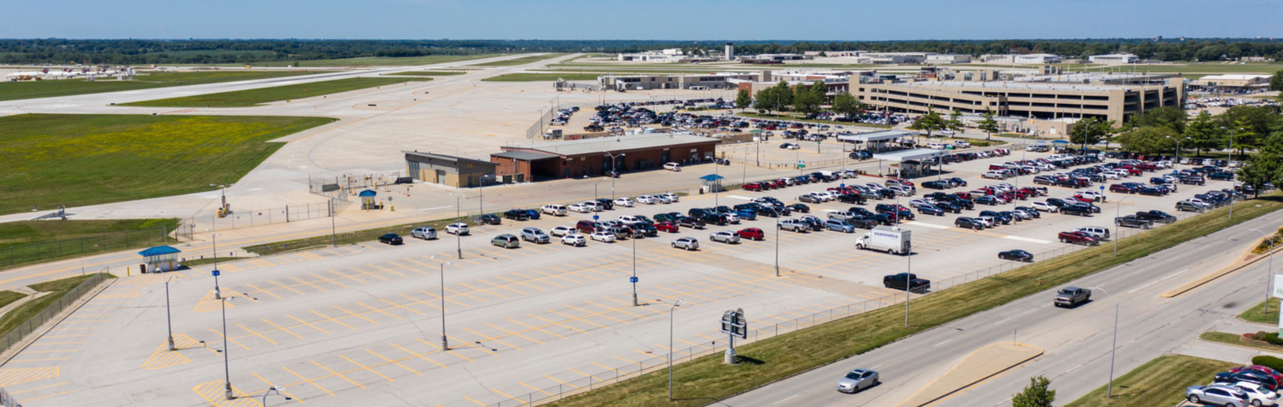 Aerial view of airport landside infrastructure including parking lots, roadways, terminal access areas, and adjacent runway facilities.