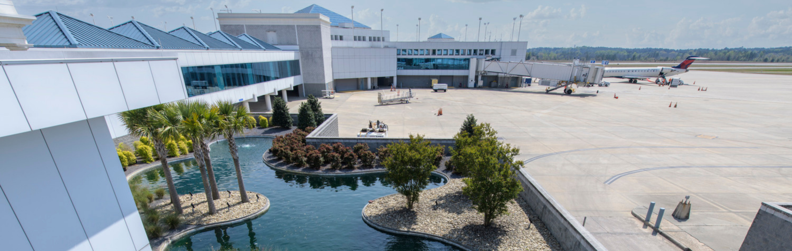 Airport terminal area with landscaped water feature, surrounding buildings, and adjacent aircraft apron, illustrating integrated aviation facility design.