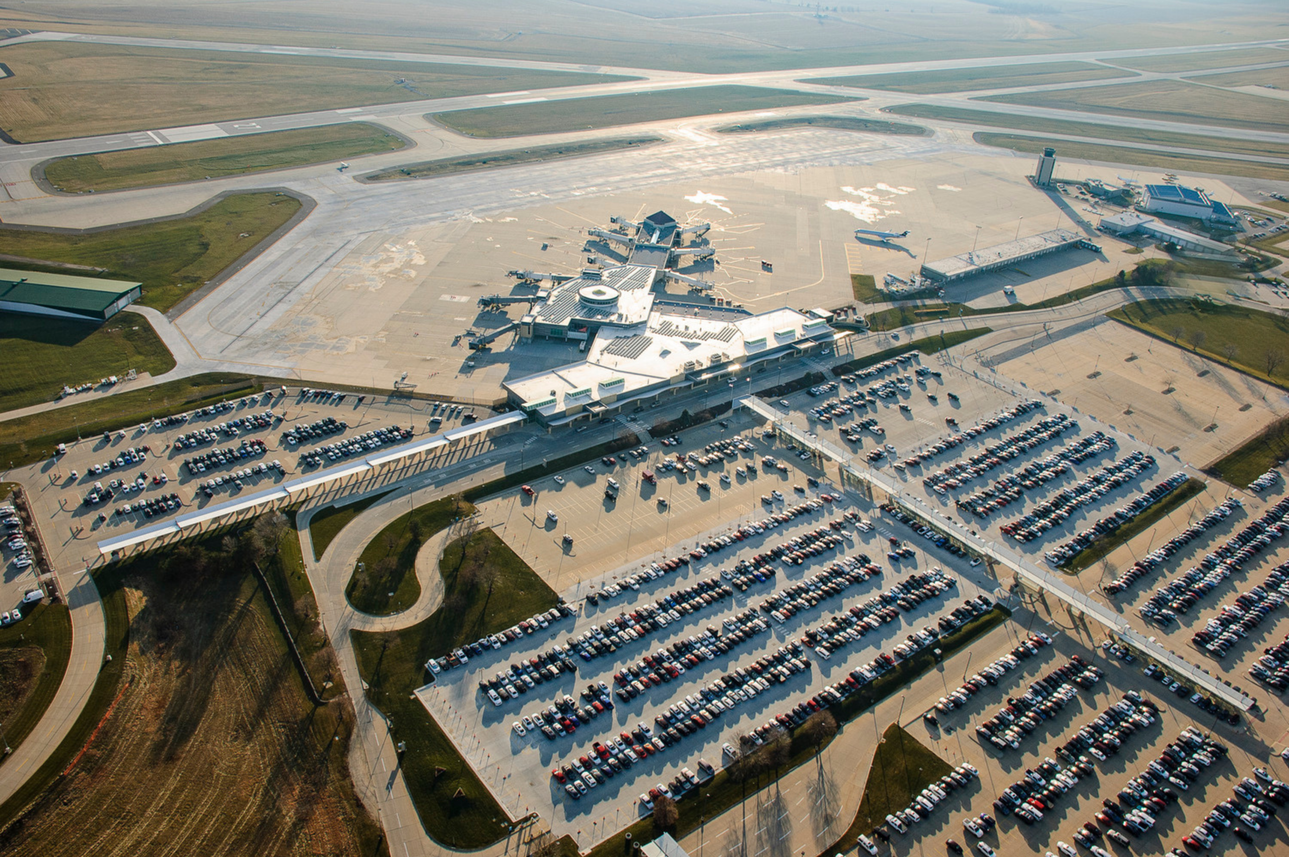 Aerial view of airport landside facilities including terminal building, parking lots, roadway access, and pedestrian connections to the airfield.